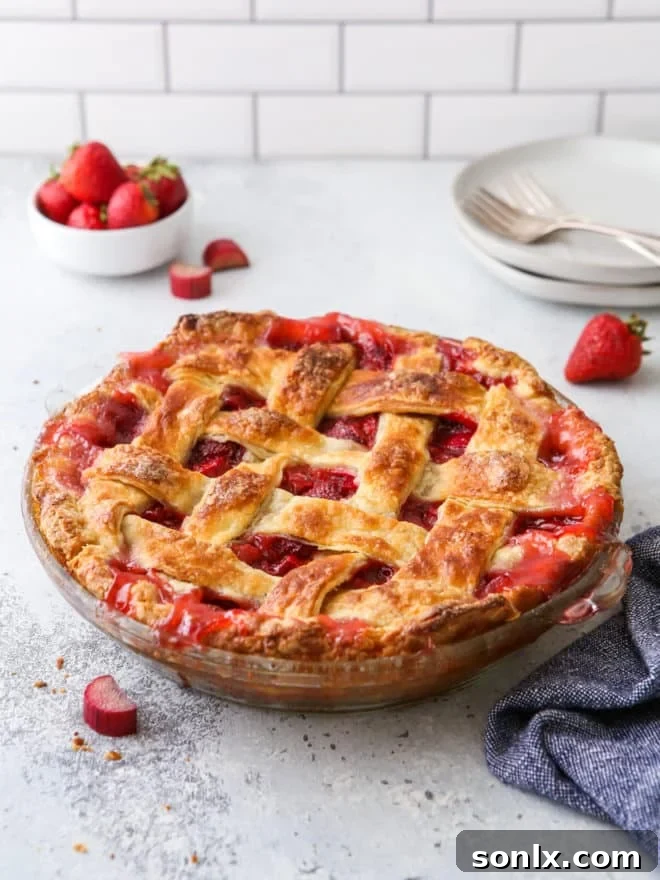 A vibrant strawberry rhubarb pie, freshly baked, on a rustic table with a stack of white plates.