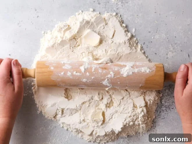 A baker using a rolling pin to flatten and integrate butter into dry ingredients on a work surface.
