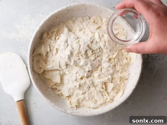 A baker gently adding ice water to the flour and butter mixture in a bowl, preparing to form the dough.