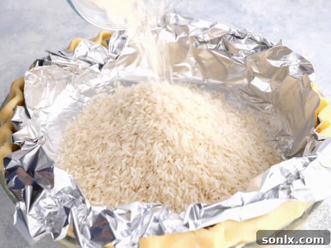 Adding dried rice as pie weights into a pie crust lined with foil, ensuring full coverage to prevent shrinking.