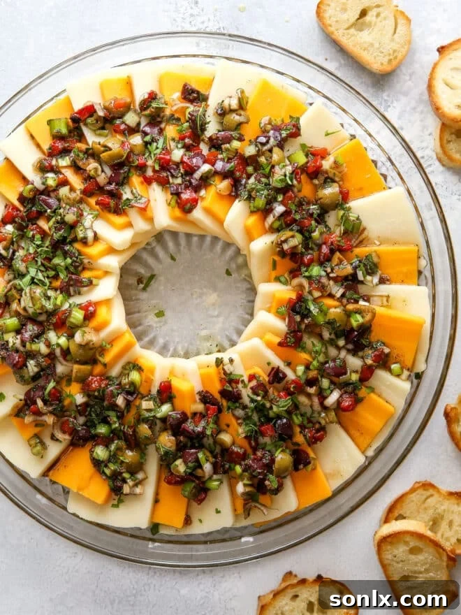 A festive marinated cheese wreath appetizer, garnished with olives, roasted red peppers, and herbs.