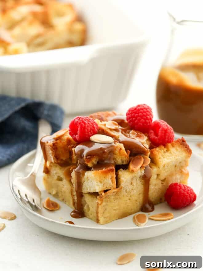 A slice of almond bread pudding on a white plate, drizzled generously with amaretto caramel sauce and garnished with fresh raspberries.