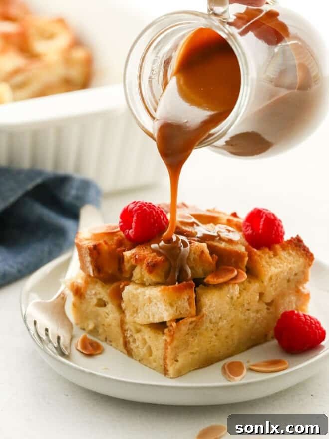 A close-up shot of a hand drizzling rich amaretto caramel sauce over a slice of almond bread pudding on a plate.