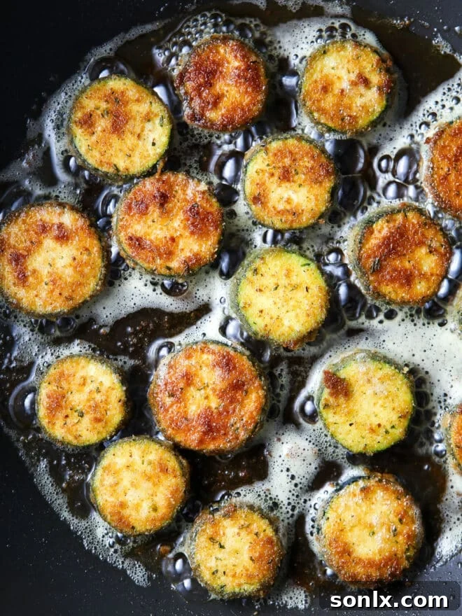 Several breaded zucchini slices sizzling and turning golden brown in a hot pan filled with oil during the frying process.