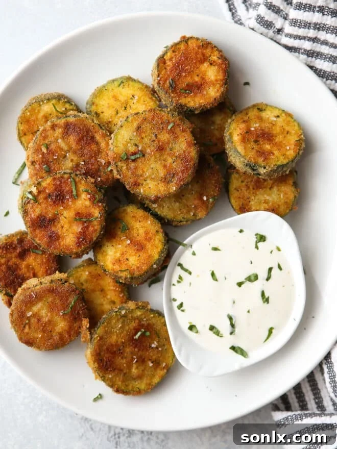 A plate of perfectly crispy golden-brown zucchini chips, garnished with fresh herbs, alongside a small bowl of ranch dressing for dipping.