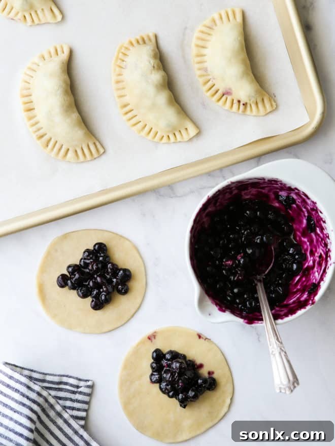 A person is carefully spooning vibrant blueberry filling onto circular pieces of pie dough on a baking sheet, preparing them for folding.