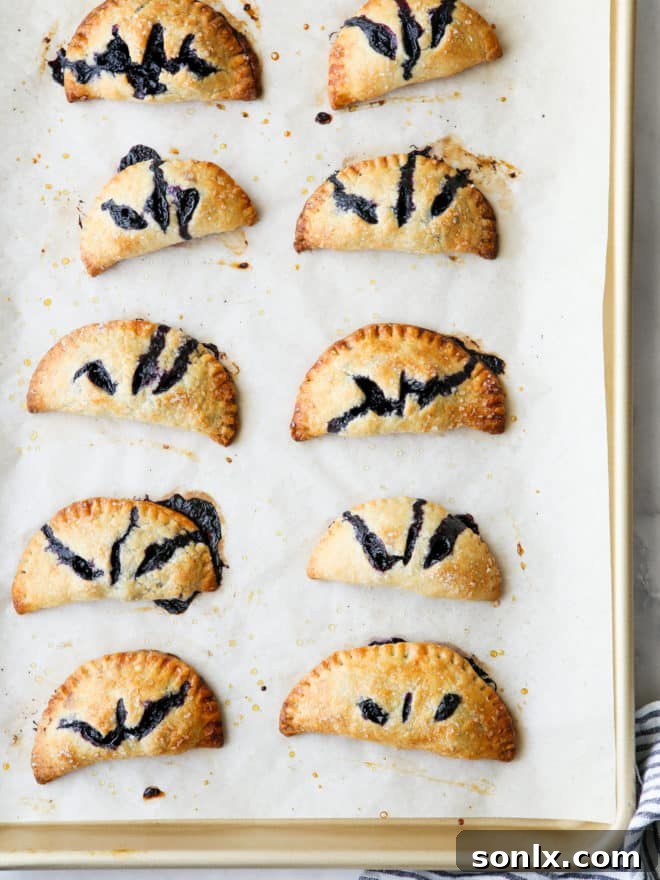 A baking sheet filled with freshly baked, golden-brown blueberry hand pies, glistening with a coarse sugar topping.