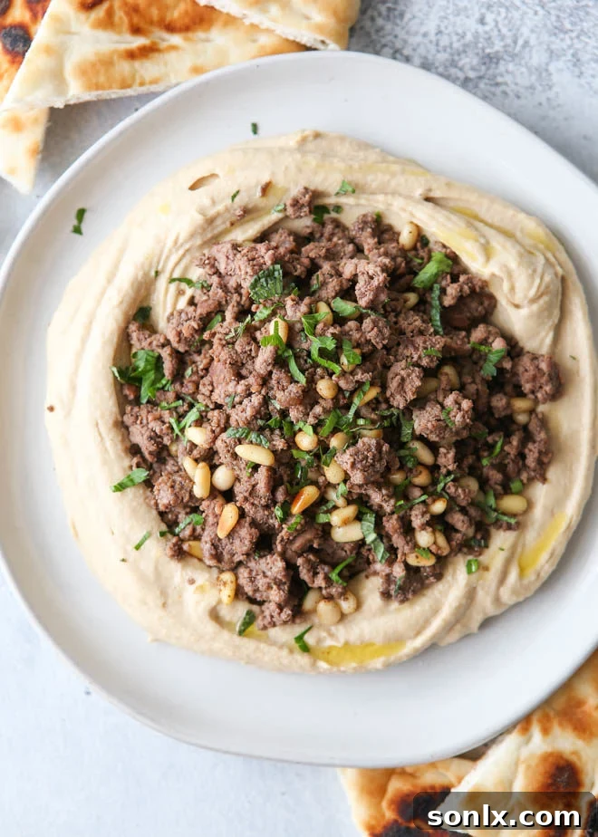 Creamy hummus topped generously with savory spiced ground beef, golden-brown toasted pine nuts, and a vibrant garnish of fresh parsley and mint, invitingly drizzled with olive oil.