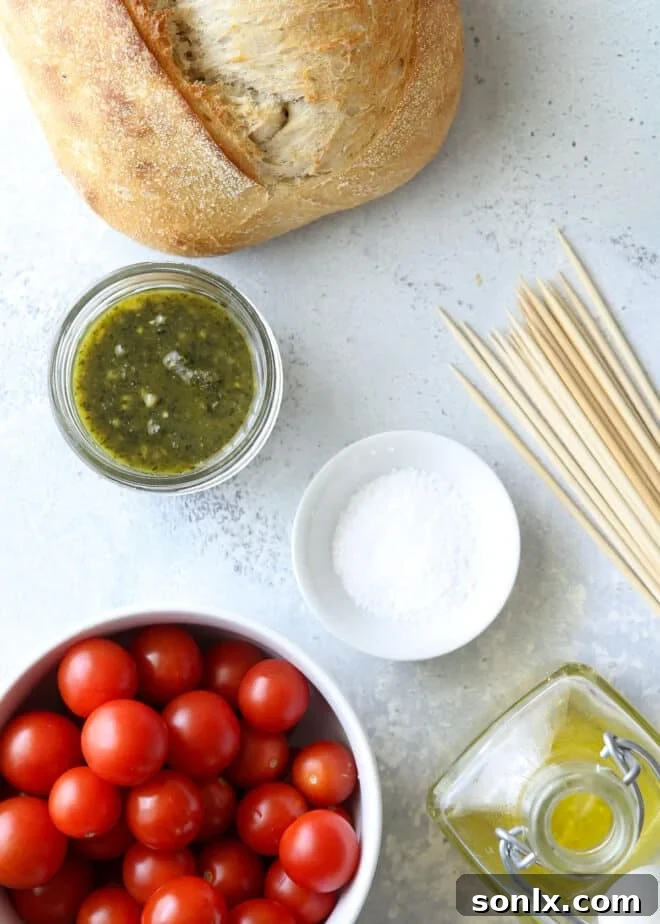 A flat lay photograph showcasing the simple yet essential ingredients needed for grilled bruschetta skewers: a crusty loaf of bread, cherry tomatoes, a jar of pesto, olive oil, and coarse salt.