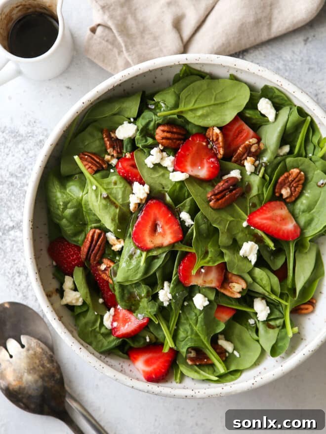 A top-down view of a generously sized bowl of strawberry spinach salad, garnished with extra ingredients and salad tongs for serving.
