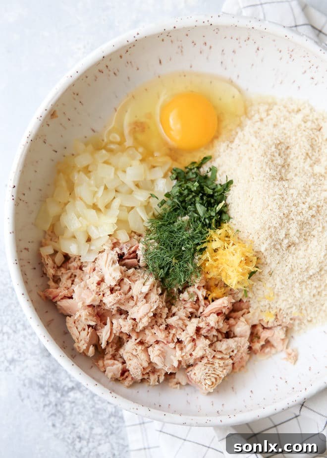 Hands mixing ingredients for salmon patties in a large bowl, showing flaked salmon, chopped herbs, and breadcrumbs.