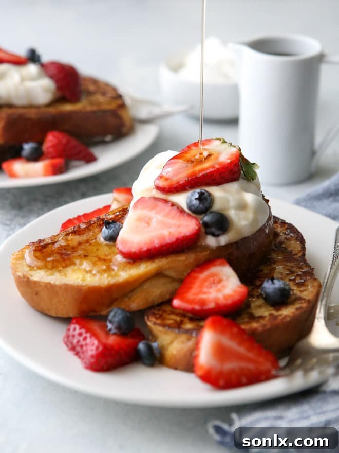 A beautifully arranged plate of French toast, generously topped with an assortment of fresh berries, a dollop of whipped cream, and a rich pour of maple syrup.