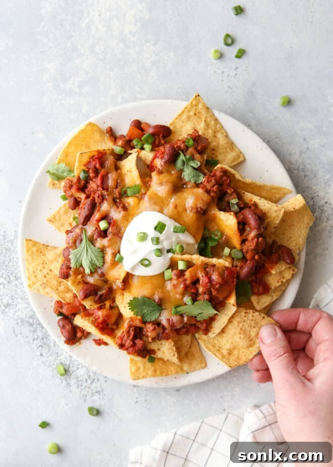 A hand reaching for a crispy chip from a plate of delicious chili cheese nachos.