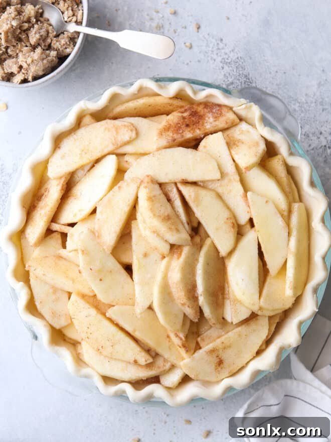 Prepared apple slices nestled in a pie crust, with the pecan and oat crumble topping resting in a separate bowl beside it.