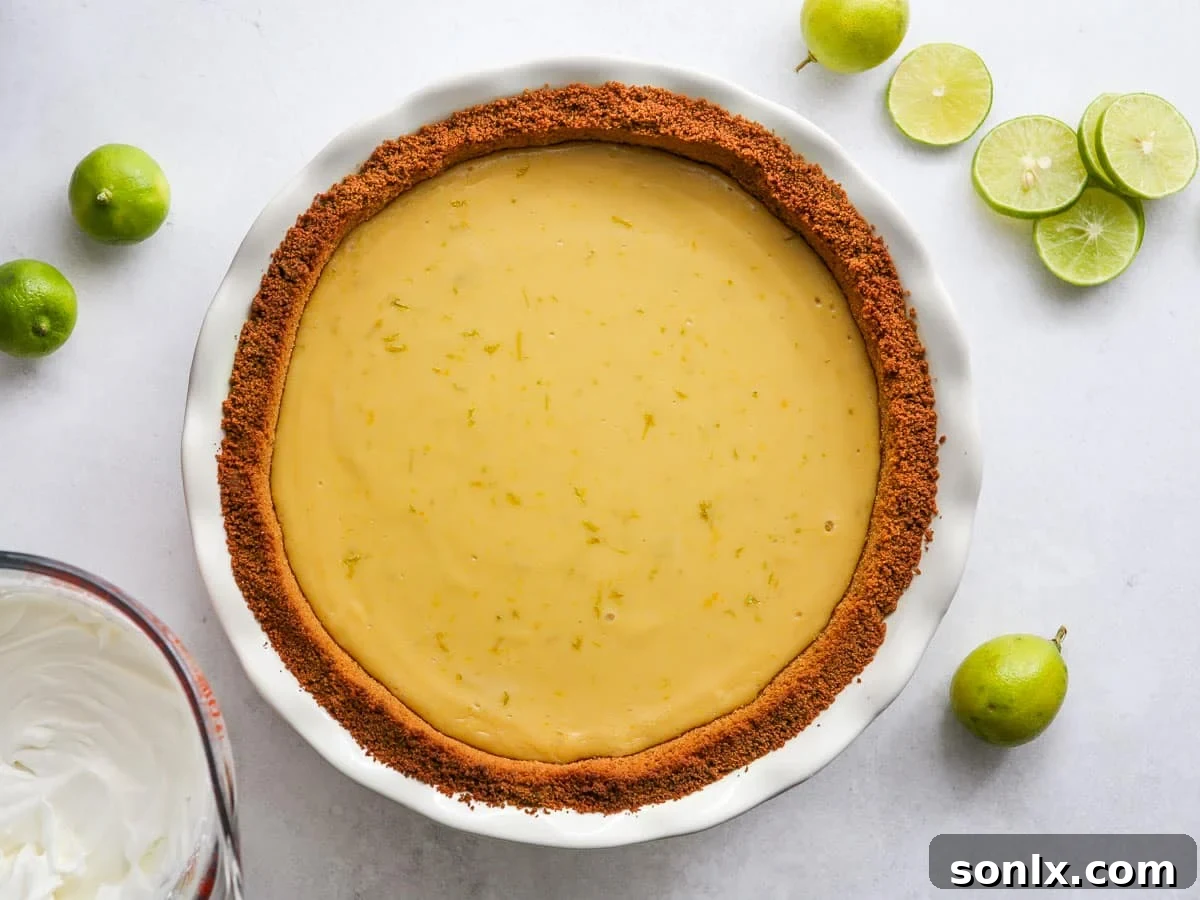 A freshly baked key lime pie cooling in a pie plate, with a bowl of whipped cream and sliced limes nearby, ready for garnishing.