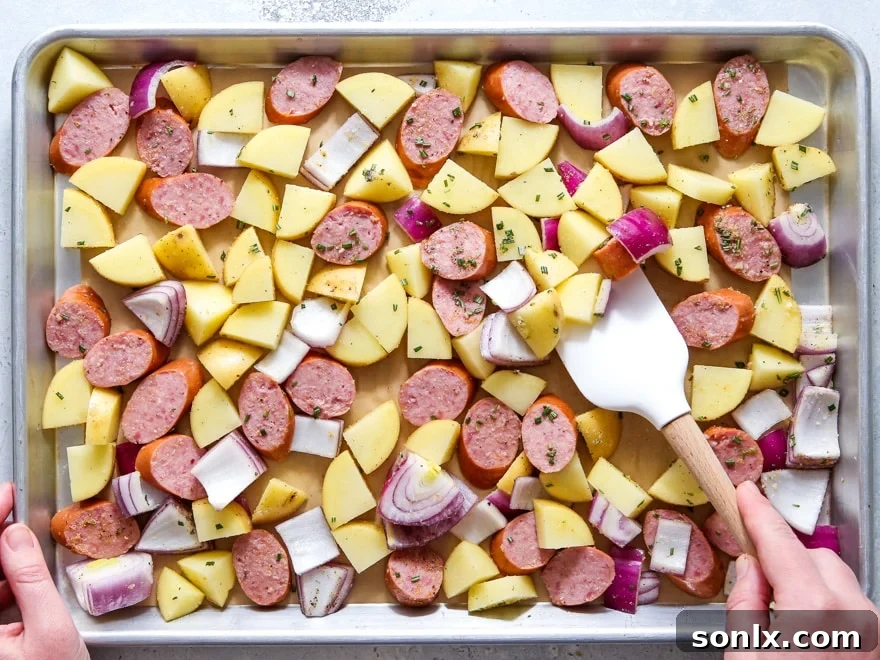 A sheet pan loaded with seasoned sausage, potatoes, and onions, spread in a single layer, ready to be baked.