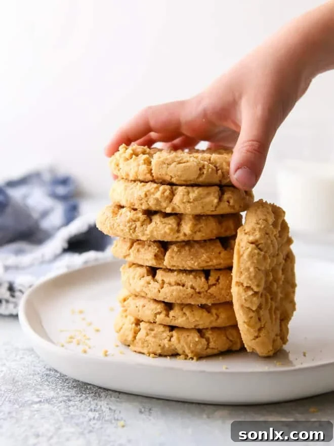 A hand reaching for a perfectly baked flourless peanut butter cookie from a stack on a plate.