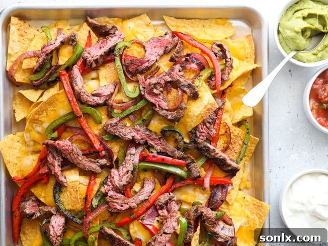 A close-up of a loaded plate of steak fajita nachos, featuring melted cheese, steak, peppers, onions, and generous dollops of guacamole, sour cream, and salsa.
