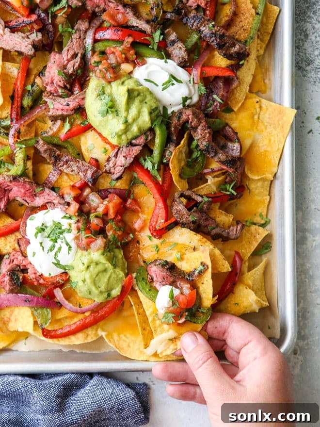 A hand reaching for a bite of a perfectly loaded steak fajita nacho from a sheet pan, highlighting the delicious layers of ingredients.