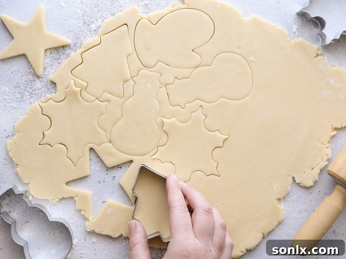 A baker using festive Christmas cookie cutters to cut out shapes from rolled sugar cookie dough on a lightly floured surface.