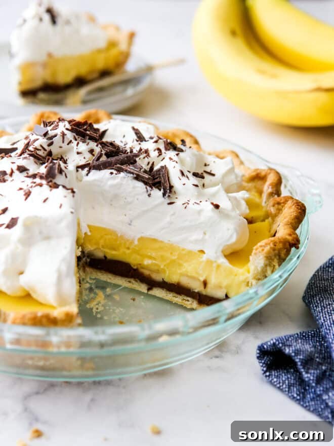 A slice of Chocolate Banana Cream Pie removed from the whole pie, showcasing its beautiful layers of crust, chocolate ganache, banana, vanilla pudding, and whipped cream, ready to be enjoyed.