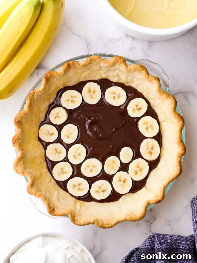 Hands carefully assembling the layers of chocolate banana cream pie in the baked crust, showing the ganache, banana slices, and vanilla pudding before the final whipped cream topping.