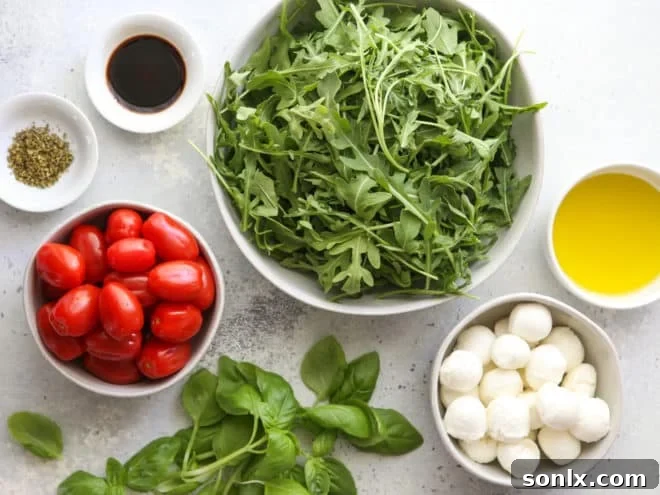 Fresh ingredients for Arugula Caprese Salad laid out on a wooden board: cherry tomatoes, mini mozzarella balls, fresh basil leaves, and a bunch of arugula.