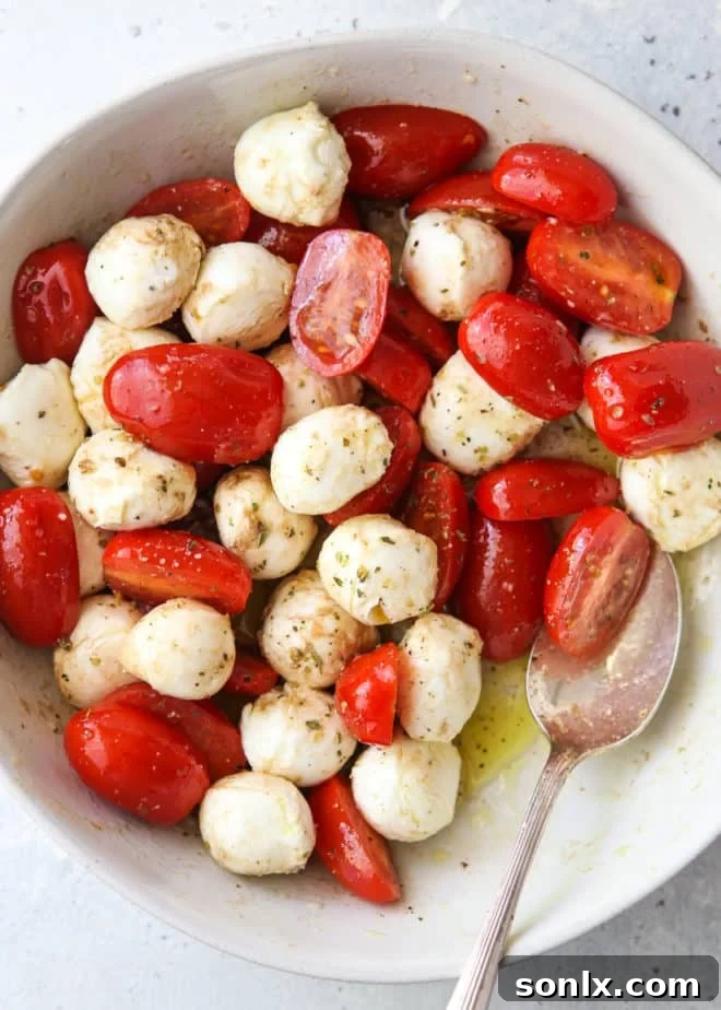 Close-up of halved cherry tomatoes and mini mozzarella balls marinating in olive oil, balsamic vinegar, and Italian seasoning in a glass bowl.