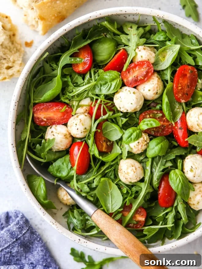 A vibrant Arugula Caprese Salad, showcasing glossy tomatoes, white mozzarella, green basil, and dark arugula, in a large serving bowl.