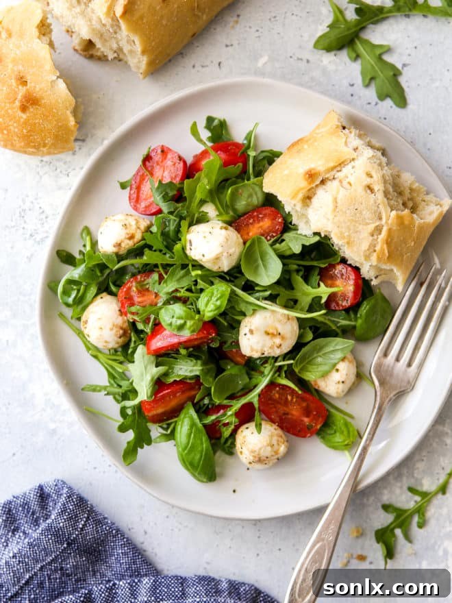 A serving of Arugula Caprese Salad on a white plate with a slice of toasted bread, emphasizing its delightful presentation.