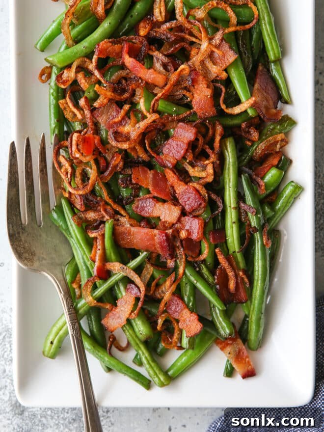 Closeup of vibrant green beans topped with golden crispy shallots and savory bacon bits, served on a elegant platter with a fork ready to enjoy.
