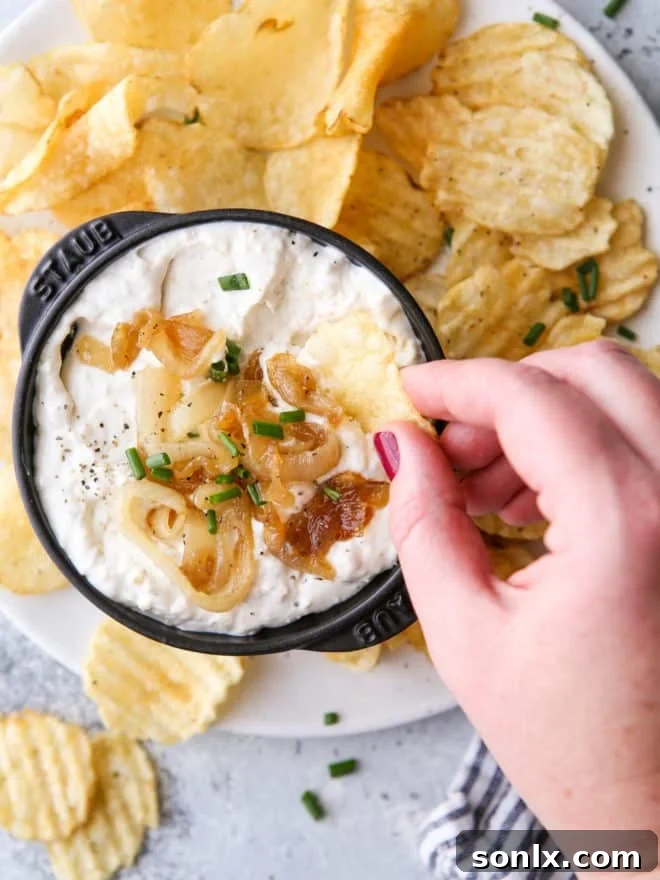 Rich Caramelized Onion Dip 7 A hand dipping a potato chip into a bowl of homemade French onion dip, showing its creamy texture.