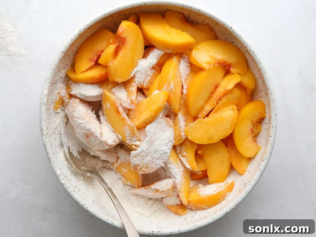 A large bowl filled with sliced peaches, sugar, flour, and cinnamon, being gently mixed for the pie filling.