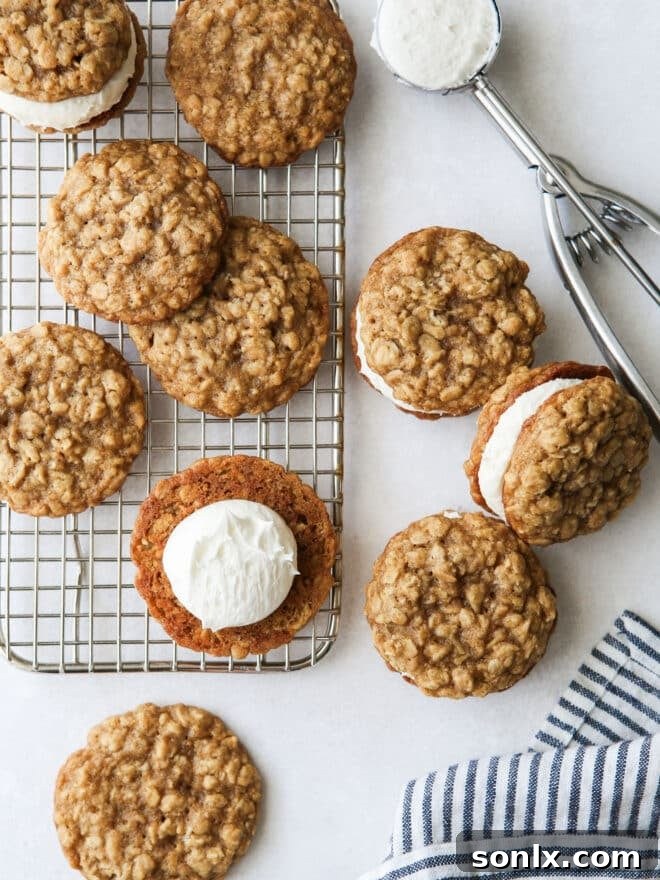 Hands assembling an oatmeal cream pie cookie by placing marshmallow filling onto one cookie and topping it with another, with other cookies cooling on a rack in the background.