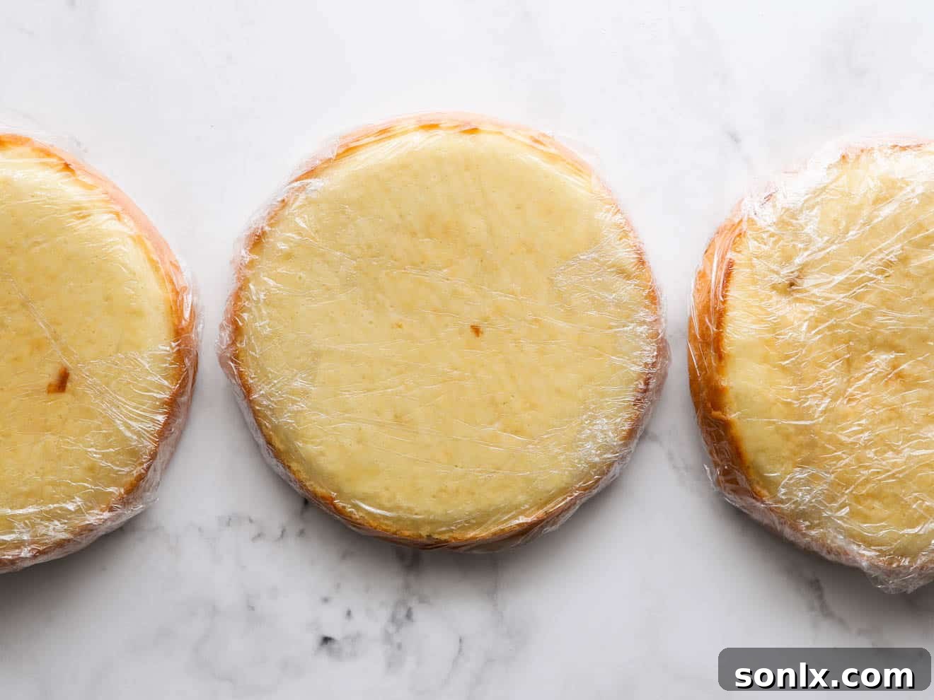 Individual coconut cake layers, perfectly golden and tender, wrapped tightly in plastic wrap, ready for chilling.