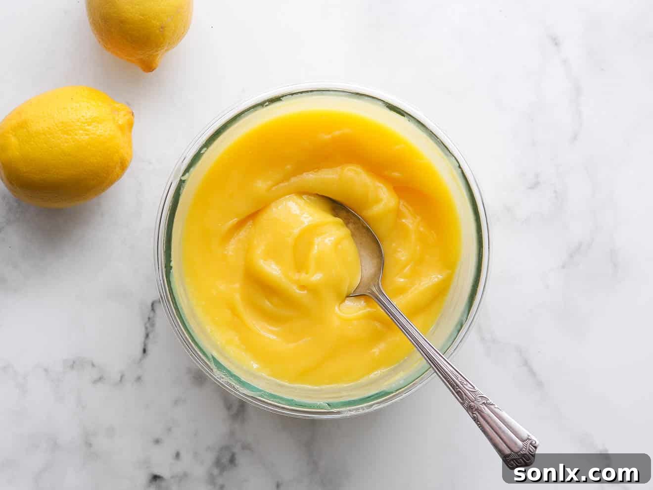 A glistening bowl of luscious homemade lemon curd, with a spoon resting in its rich, smooth surface, ready to be chilled.