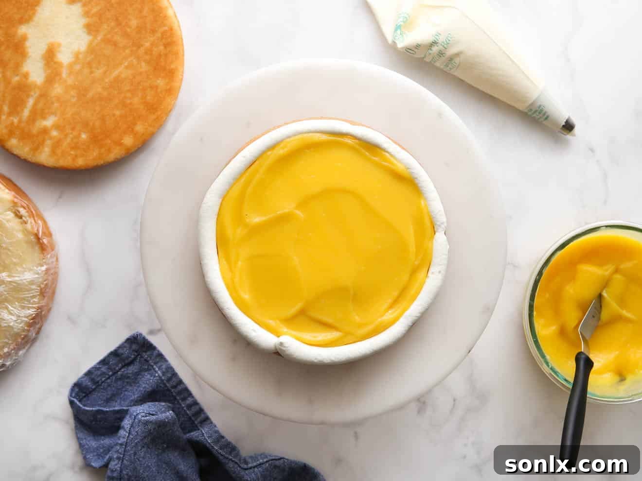 A cake layer being filled with bright yellow lemon curd, surrounded by a piped dam of white Chantilly frosting, on a cake stand.