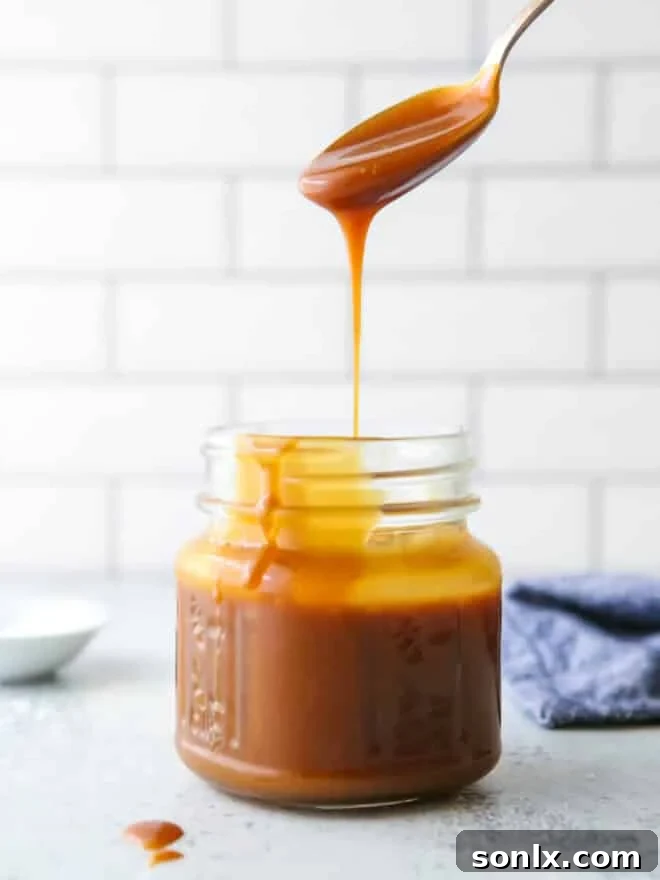 A spoonful of golden homemade caramel sauce being poured into a glass jar, highlighting its rich, glossy texture.