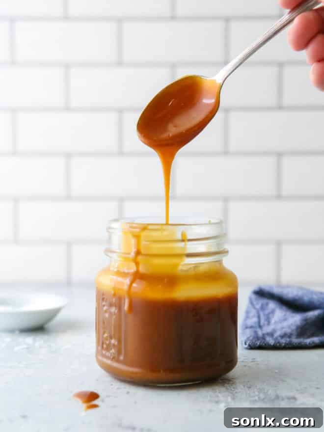 Homemade caramel sauce being poured from a spoon into a clear glass jar, showing its rich, flowing consistency.