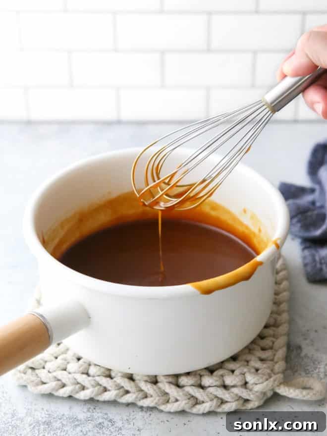 A whisk stirring smooth, shimmering homemade caramel sauce in a stainless steel pan.