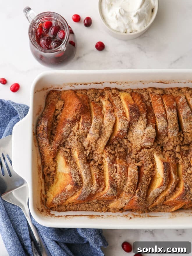 Baked French toast casserole in a baking pan, served with a pitcher of cranberry maple syrup and a bowl of whipped cream, on a festive table setting.