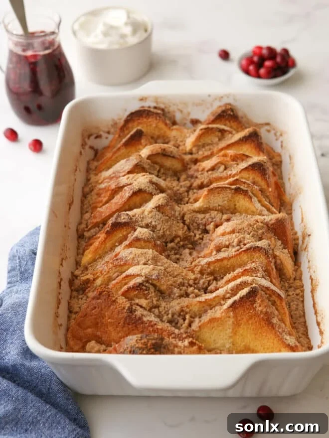 Baked French toast in a baking pan, garnished with whipped cream and a pitcher of cranberry maple syrup, ready to be served.
