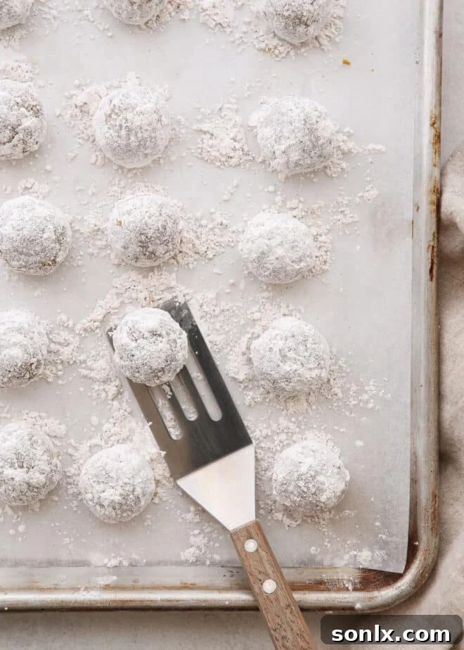 A baking sheet filled with freshly baked Molasses Spice Snowball Cookies, still warm from the oven, lightly coated in powdered sugar and ready for a second dusting.