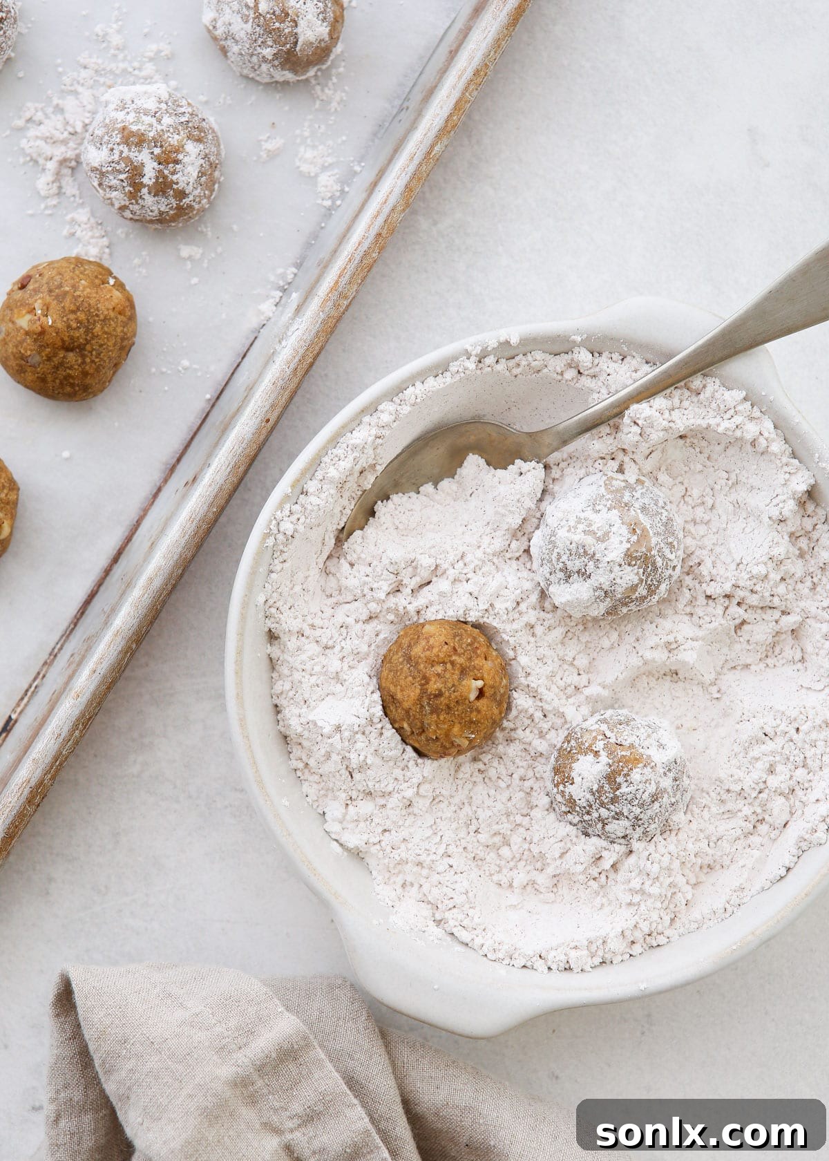 A hand gently rolling a warm, freshly baked Molasses Spice Snowball Cookie in a bowl filled with cinnamon-powdered sugar. The cookie is getting its first, light coating.