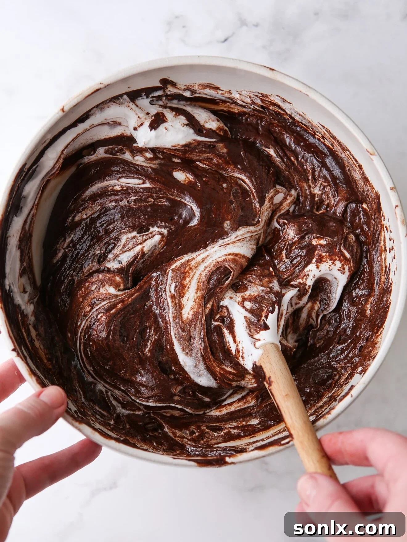 Hands gently folding rich chocolate batter, a crucial step in preparing a decadent chocolate dessert.