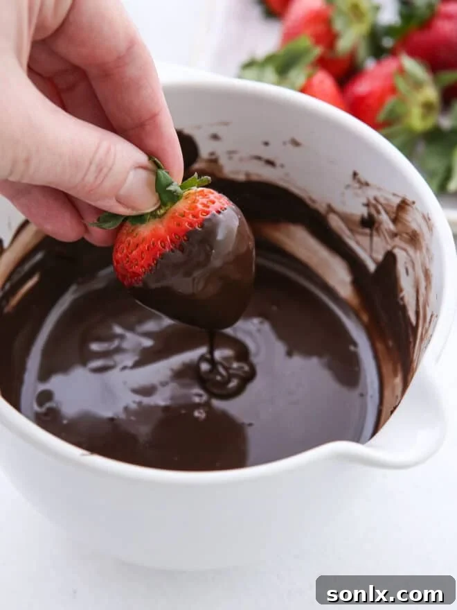 A fresh strawberry being carefully dipped into a bowl of melted, smooth chocolate.