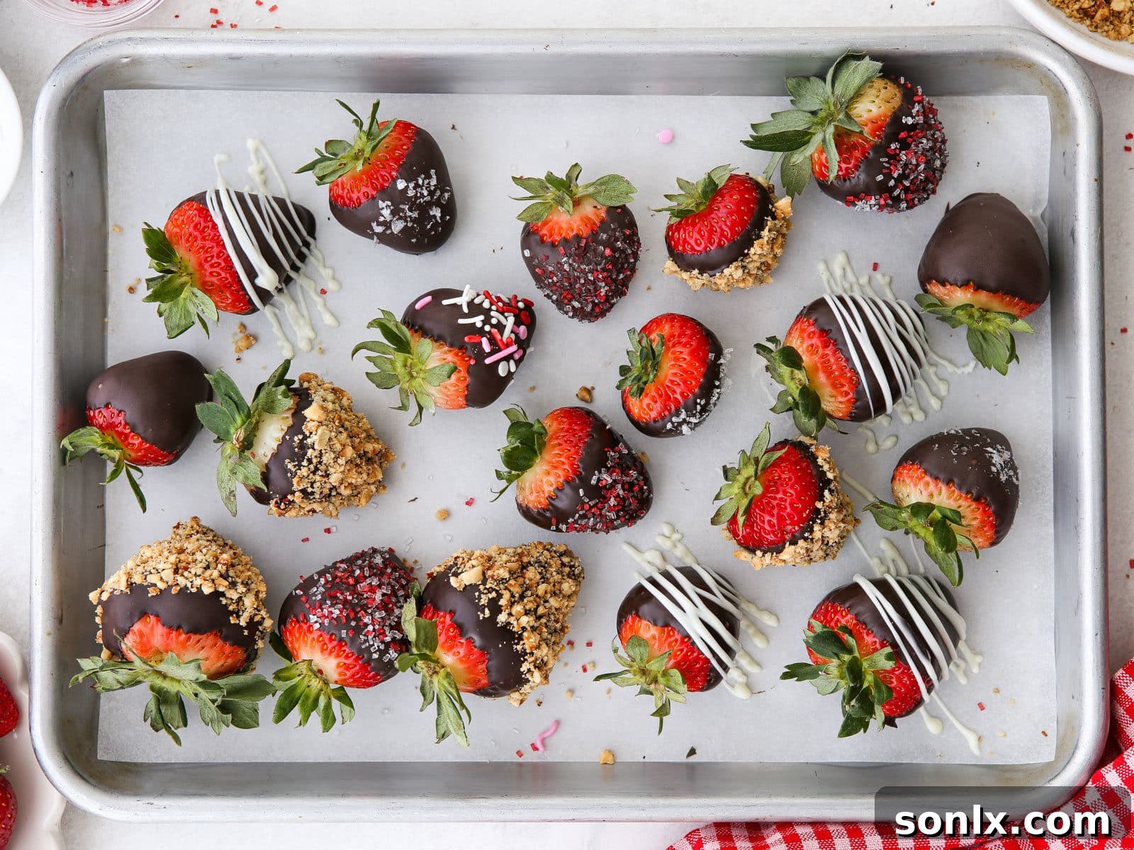 A baking sheet filled with decorated chocolate-covered strawberries, some with sprinkles, others with nuts.