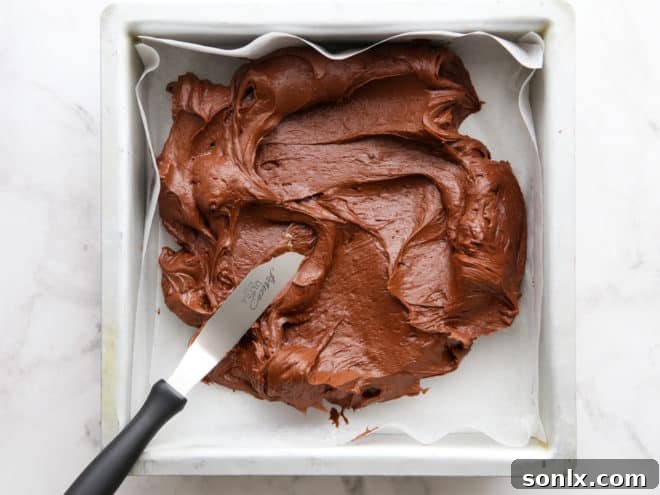 A spatula spreading freshly made peppermint fudge mixture evenly into a parchment-lined 8x8 inch baking pan, preparing it for chilling.