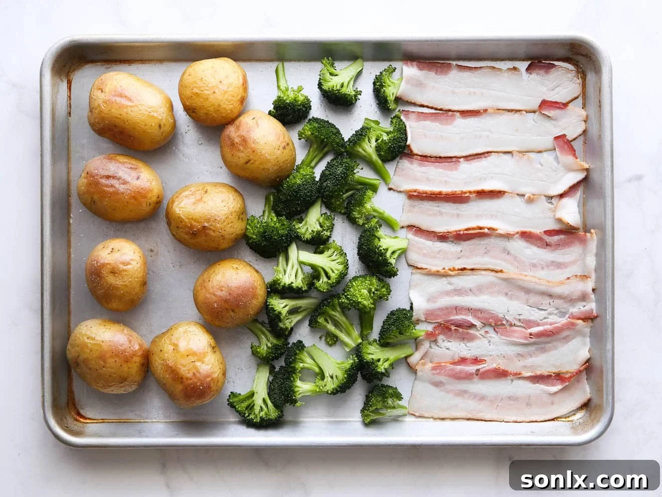 Broccoli florets and halved bacon strips being added to a sheet pan alongside partially roasted potatoes.