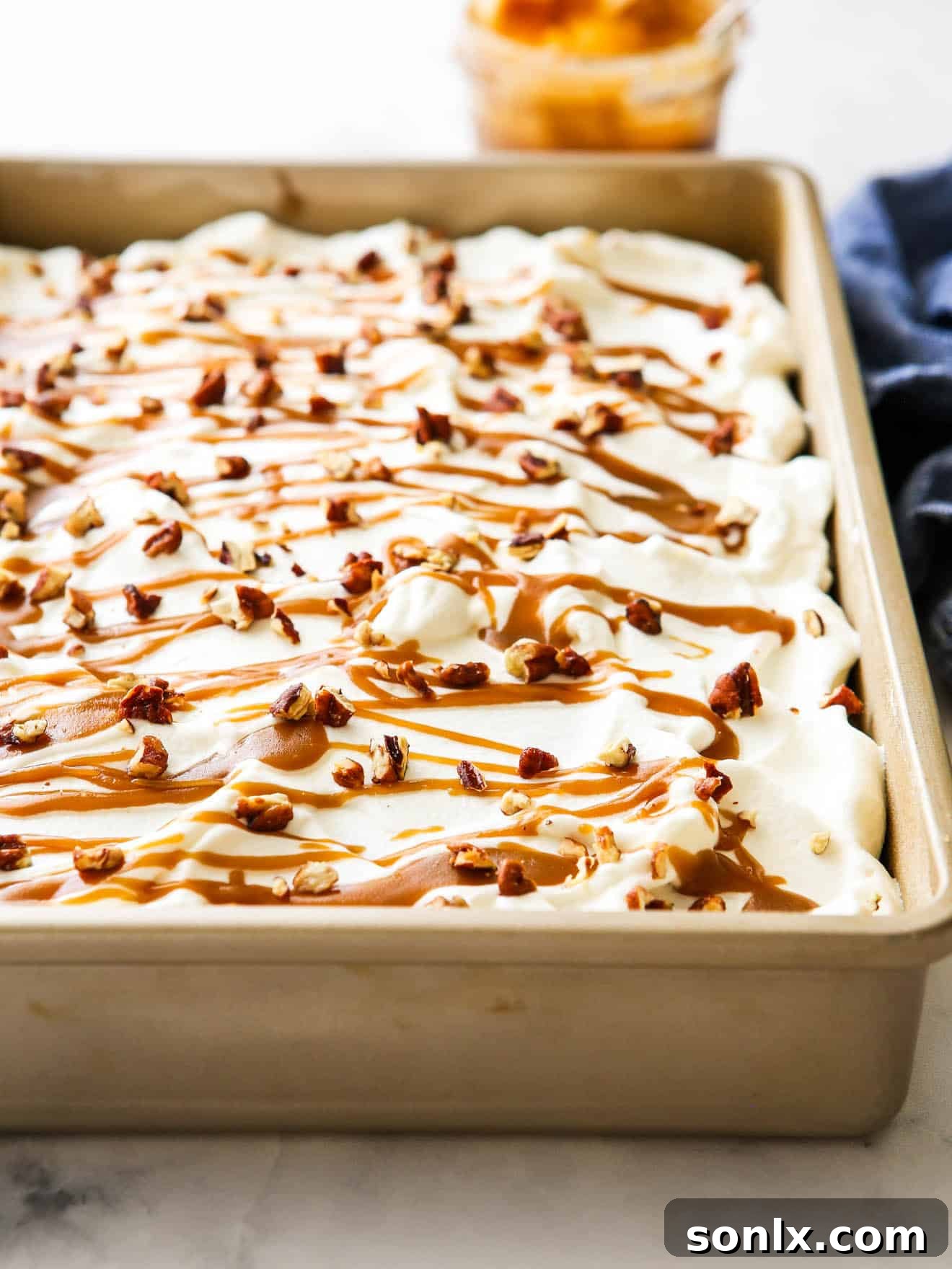 A beautifully frosted pumpkin caramel poke cake, still in its baking pan, adorned with a swirl of whipped cream, caramel drizzle, and chopped candied pecans.